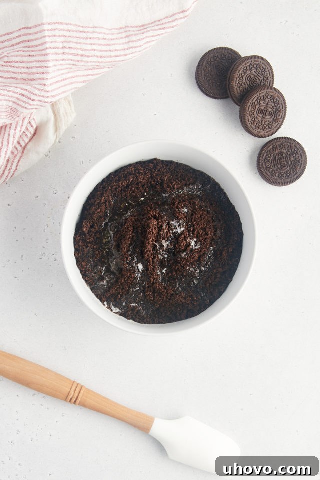 Overhead view of melted butter being added to a bowl of finely crushed Oreo cookie crumbs, ready for thorough mixing to form the cheesecake crust.