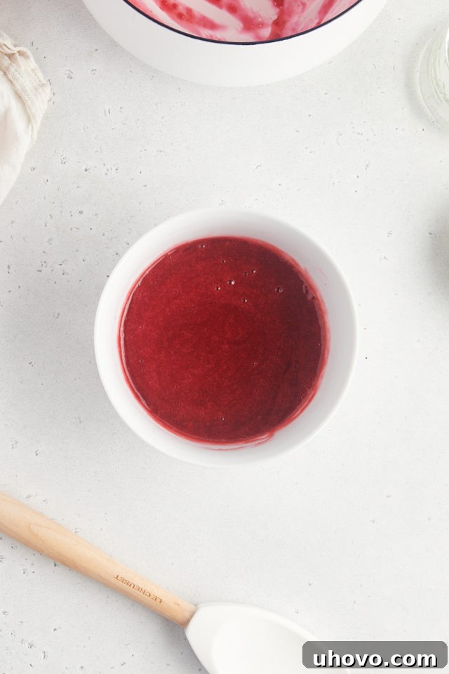 Overhead view of hot raspberry sauce being carefully strained through a fine-mesh sieve into a bowl, ensuring a perfectly smooth, seedless consistency.