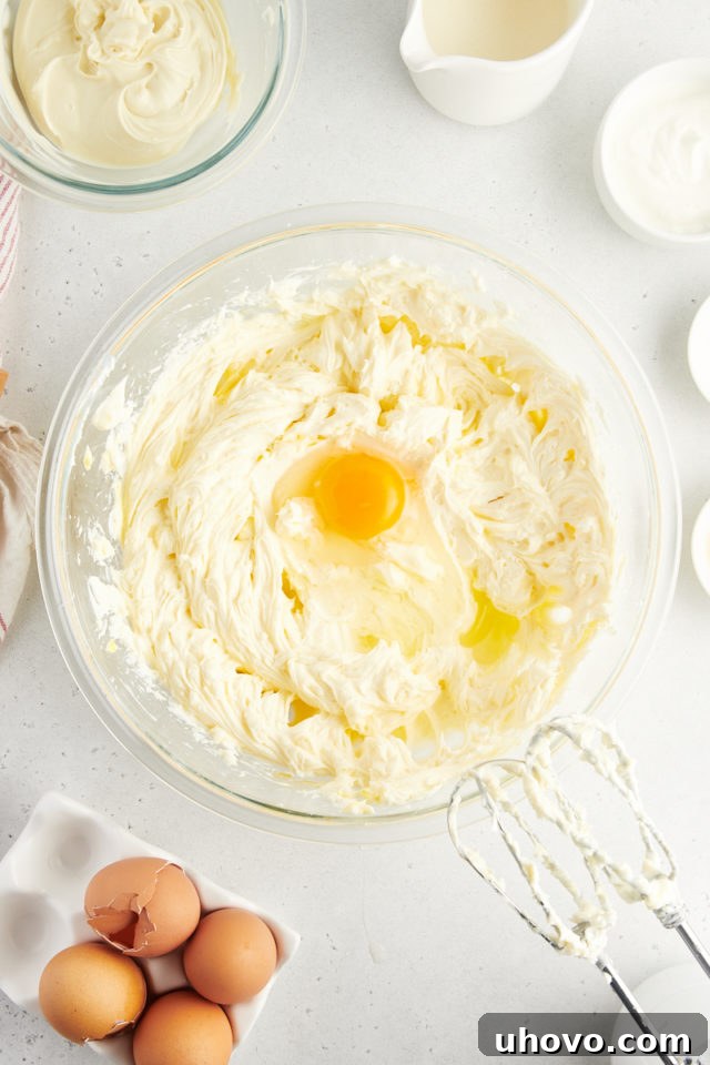 Overhead view of a single large egg being carefully added to the cream cheese mixture in a bowl, ready to be gently beaten in.