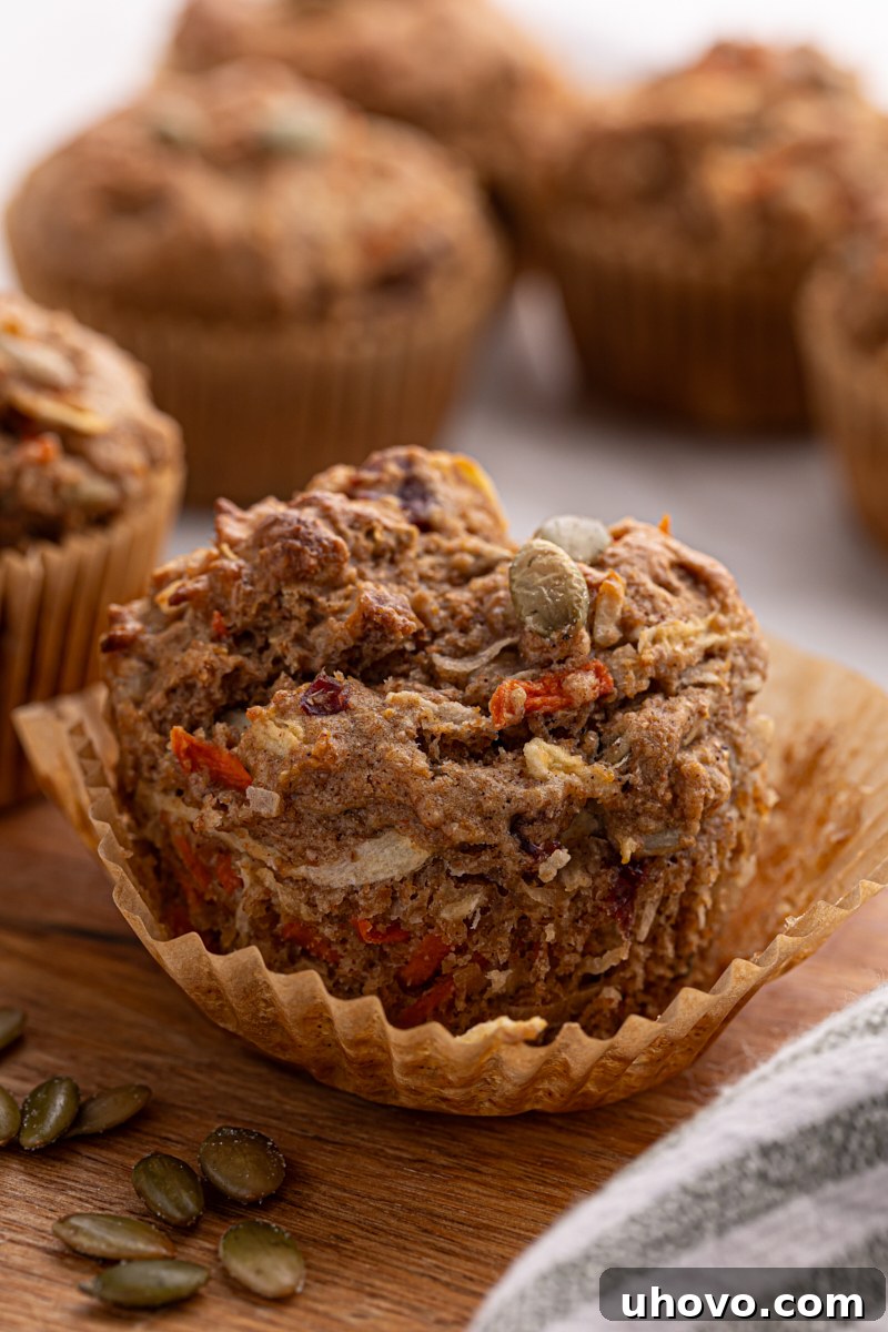 A partially unwrapped Morning Glory Muffin, showcasing its rich texture and ingredients, with more muffins blurred in the background, suggesting abundance.