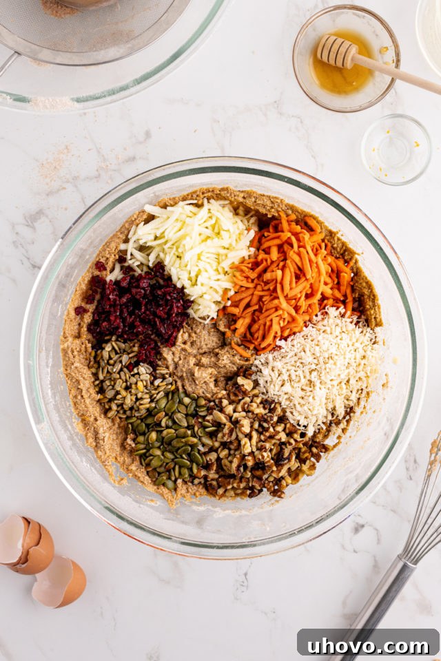 Overhead view of shredded carrots, grated apple, shredded coconut, nuts, and dried cranberries freshly added to the morning glory muffin batter in a glass mixing bowl.