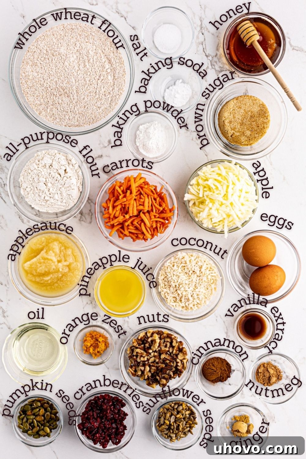 Overhead view of various fresh ingredients for Morning Glory Muffins, including grated carrots, apples, nuts, and spices laid out on a clean white surface.