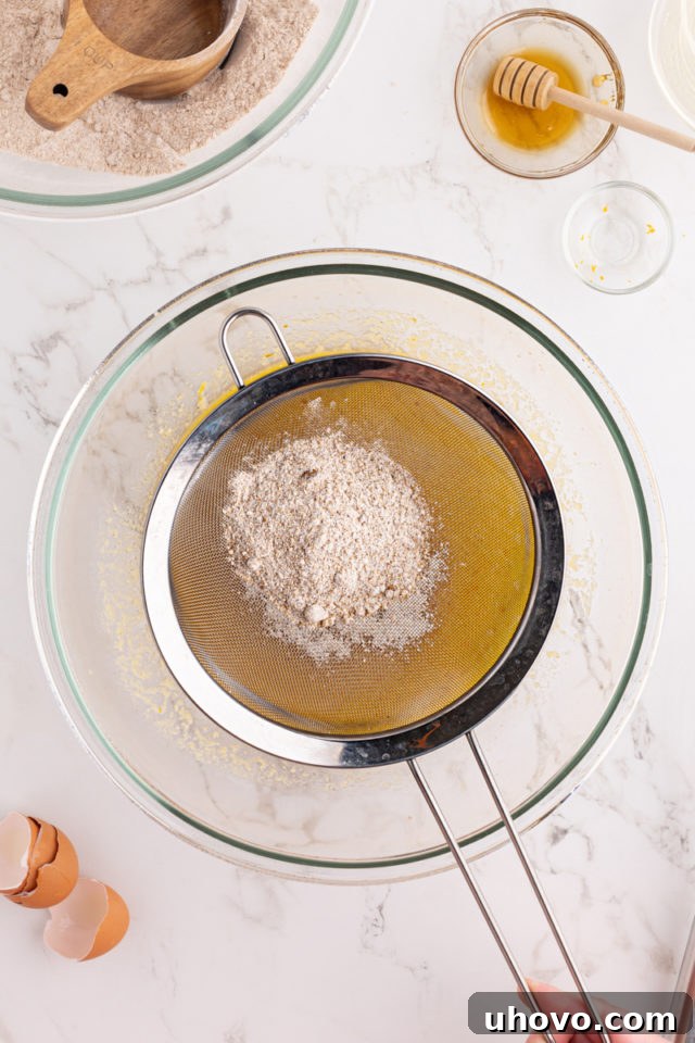 Overhead view of dry ingredients being sifted from a fine-mesh strainer into the bowl of wet ingredients for morning glory muffins, ensuring a lump-free mixture.