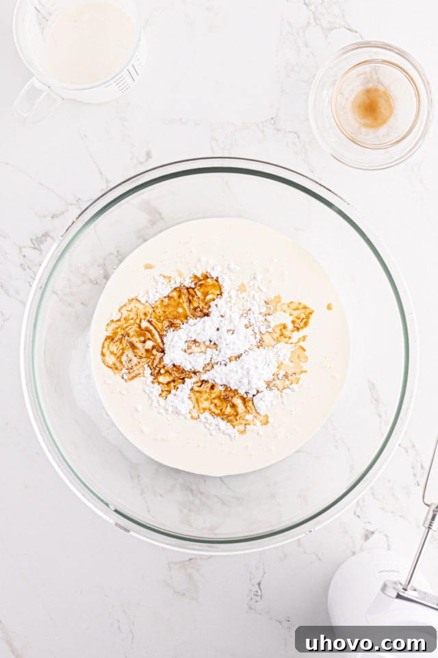 An overhead view of cold heavy cream, confectioners' sugar, and vanilla extract in a large glass mixing bowl, ready for whipping.