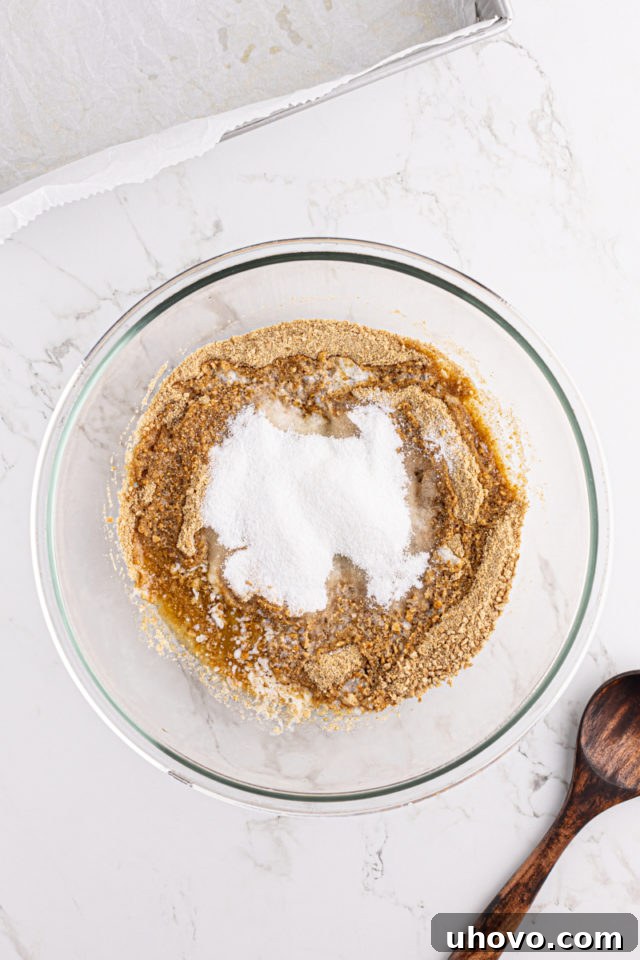 An overhead view of golden graham cracker crumbs and granulated sugar mixed together in a clear glass mixing bowl.