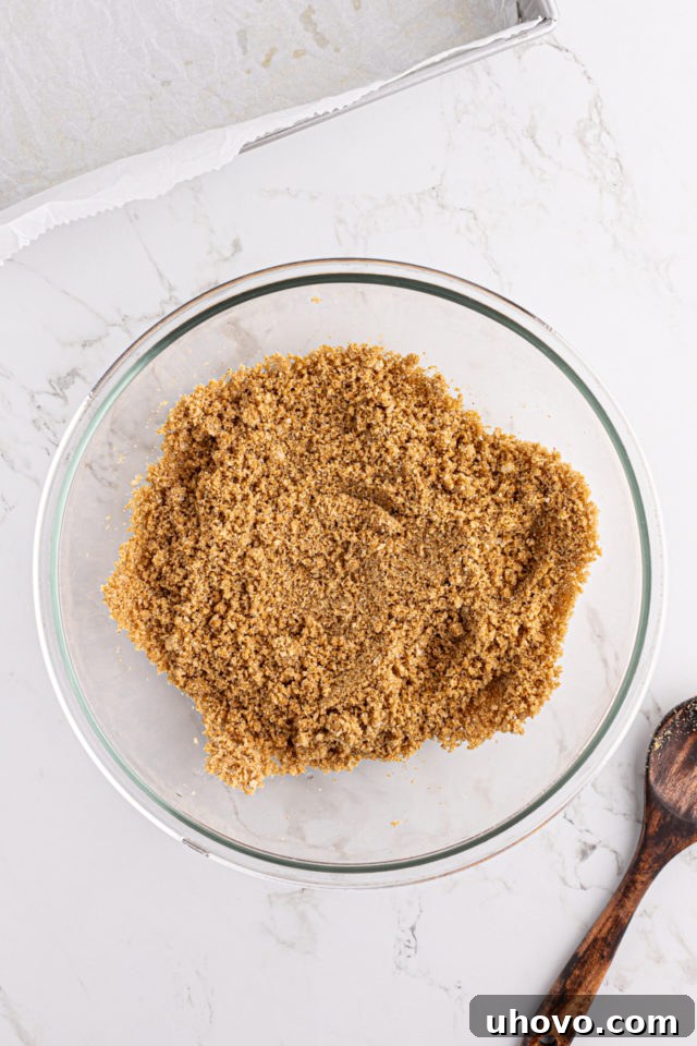 An overhead shot showing melted butter being stirred into the graham cracker and sugar mixture in a glass bowl, creating a moist, crumbly texture.