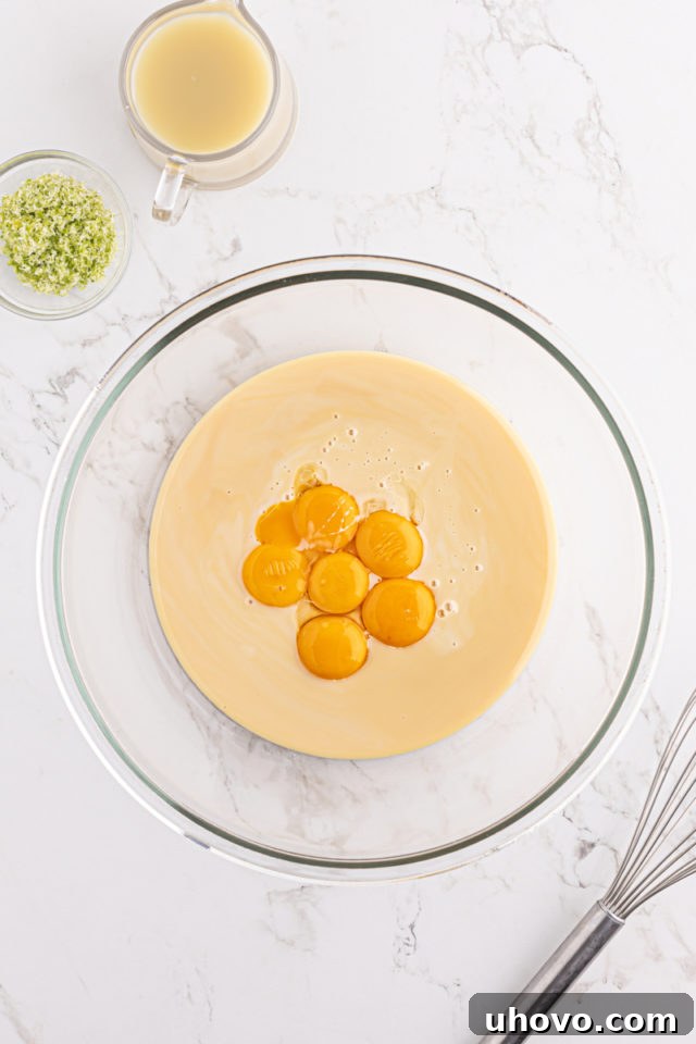 An overhead view of rich yellow egg yolks and thick sweetened condensed milk in a clear glass mixing bowl, prior to whisking.
