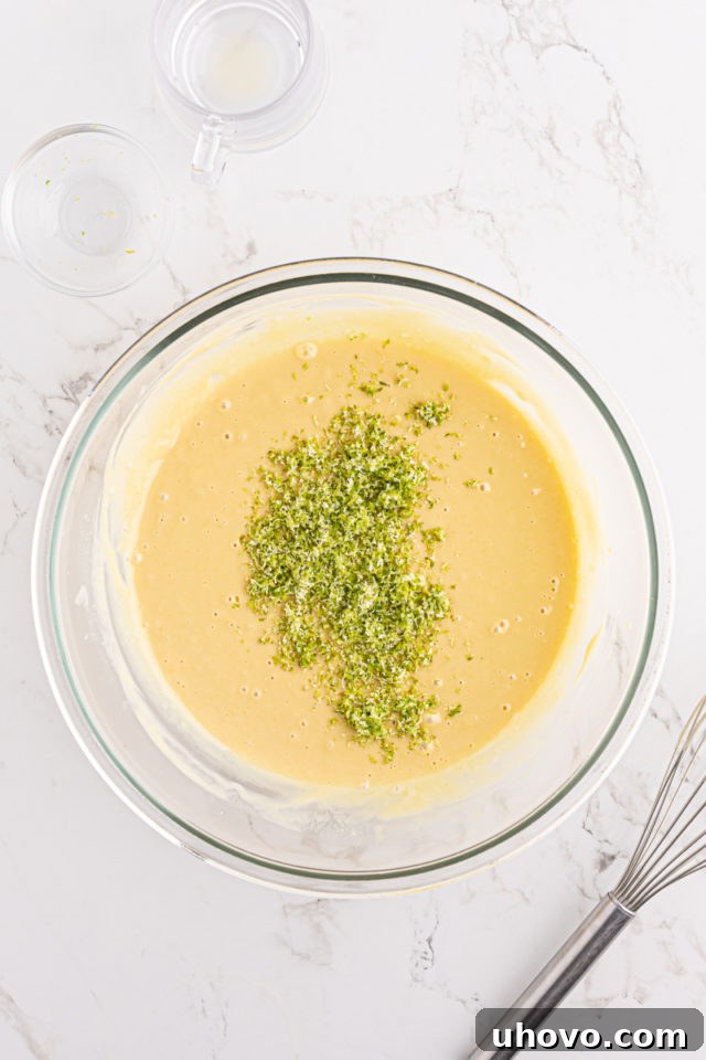 Finely grated key lime zest being added to the creamy key lime pie filling mixture in a glass bowl.