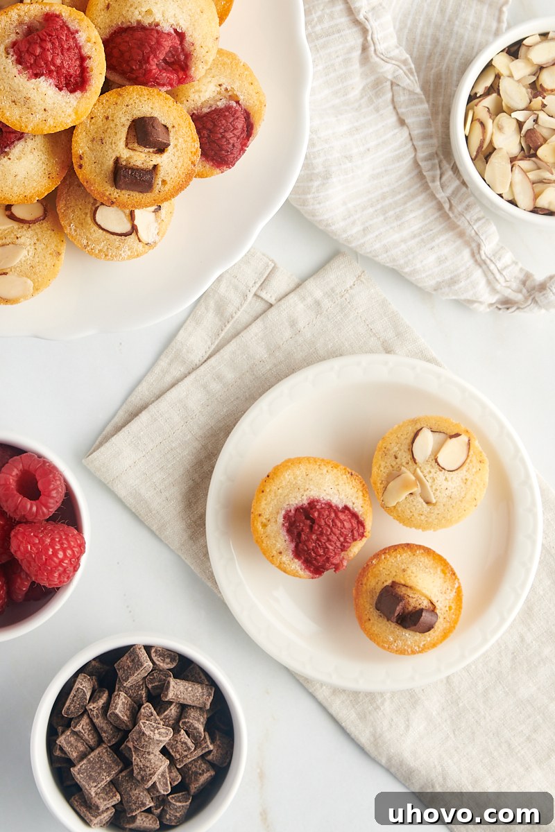 overhead view of three financiers on a white plate with more cakes and bowls of various toppings surrounding