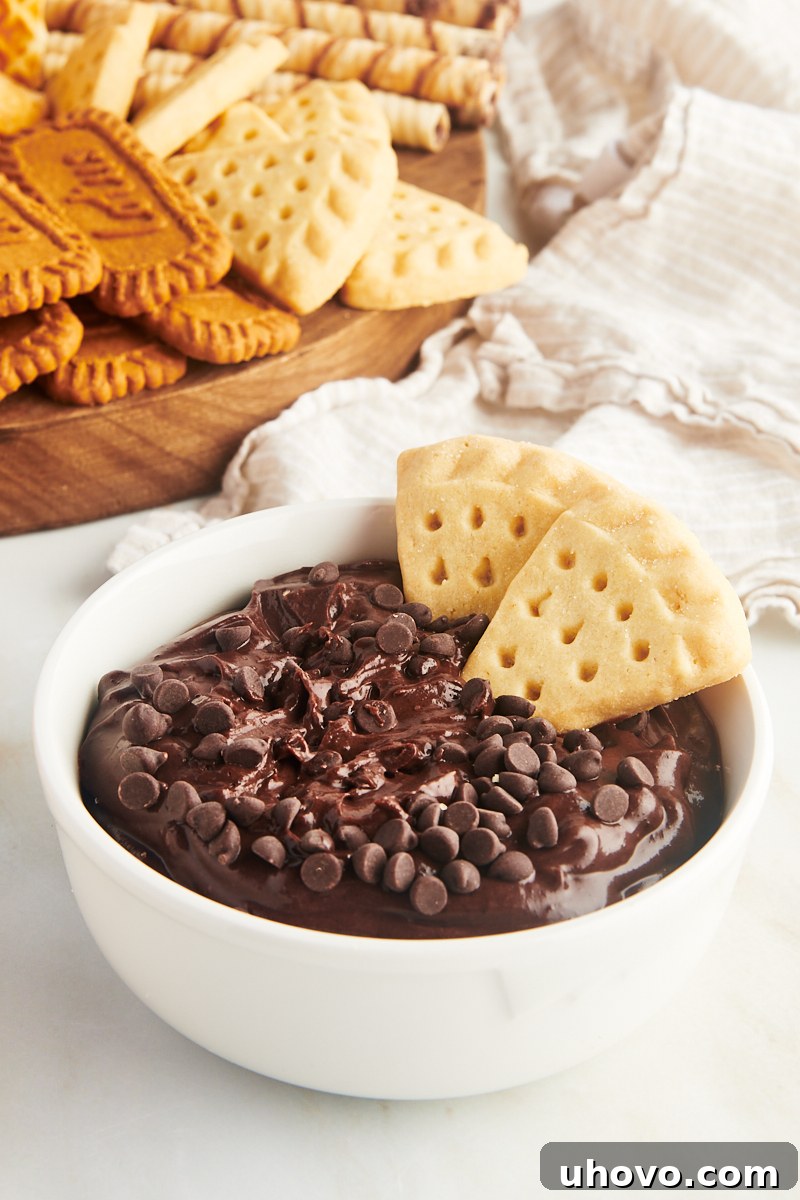 A creamy brownie batter dip in a white bowl, served with two shortbread cookies and a platter of various dippers in the background.
