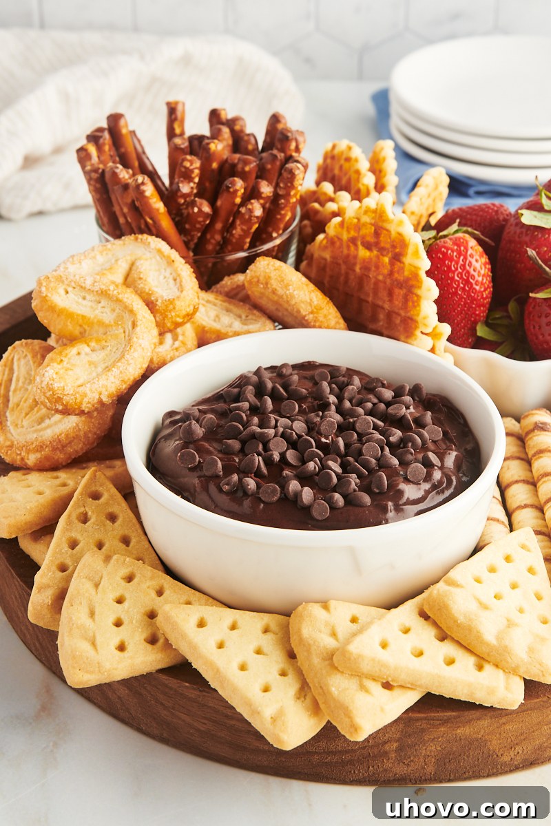 A charming wooden board displaying a central bowl of rich brownie batter dip, artfully surrounded by an inviting array of cookies, fresh berries, and crispy pretzels for dipping.