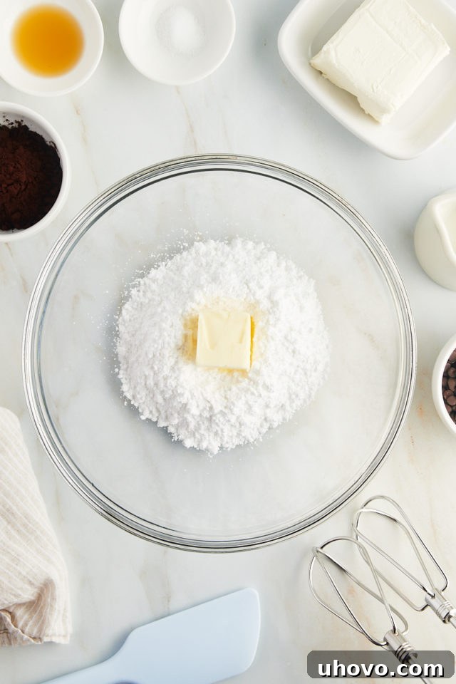 An overhead view of confectioners' sugar and softened butter being creamed together in a clear glass mixing bowl.