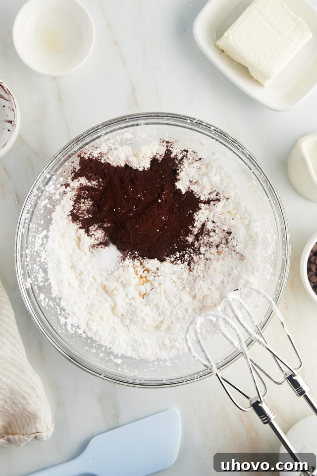 An overhead shot showing cocoa powder, vanilla extract, and a pinch of salt being added to the previously creamed butter and confectioners' sugar mixture.