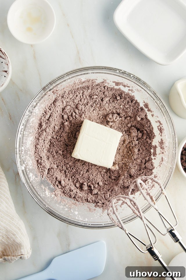 An overhead view of softened cream cheese being added to the dry ingredients for the brownie batter dip in a mixing bowl.