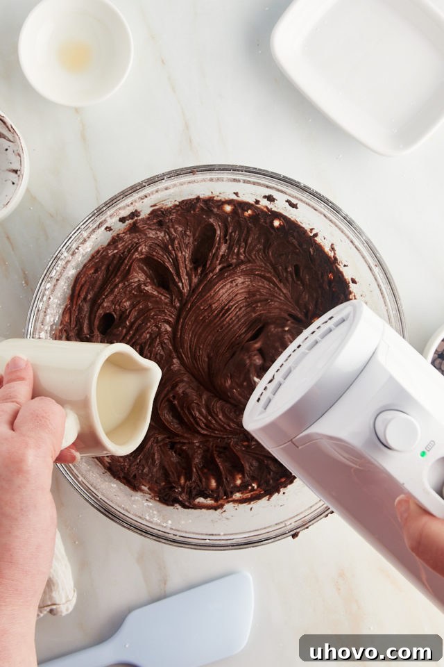 An overhead shot illustrating milk being gradually added to the brownie batter dip mixture, as it's being beaten in a glass bowl.