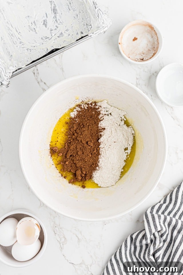Overhead view of dry ingredients, cocoa powder and flour, being added to the wet ingredients for the brownie base