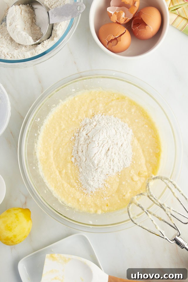 overhead view of dry ingredients being gradually added to the wet ingredients for lemon ricotta cake in a glass mixing bowl, with a spatula ready to mix