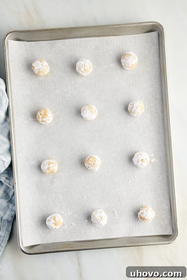 Overhead view of unbaked Amaretti cookies arranged on a parchment-lined baking sheet, spaced apart and coated in confectioners' sugar, ready for the oven.