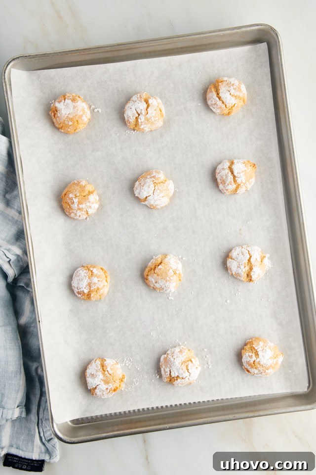 Overhead view of freshly baked Amaretti cookies cooling on a parchment-lined baking sheet, showcasing their beautifully cracked tops.