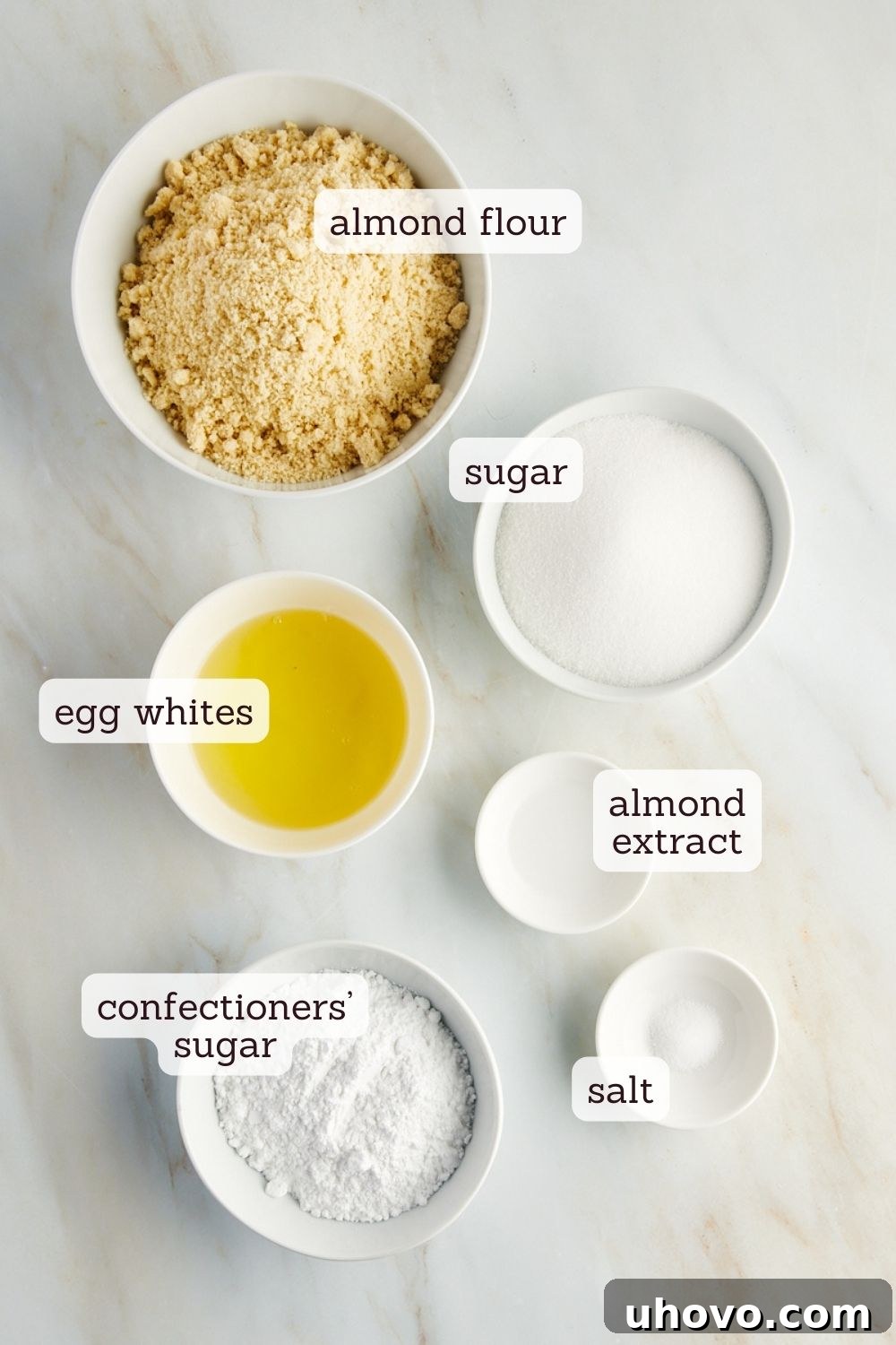 Overhead view of the six essential ingredients for Amaretti cookies, neatly laid out with labels: almond flour, granulated sugar, salt, egg whites, almond extract, and confectioners' sugar.
