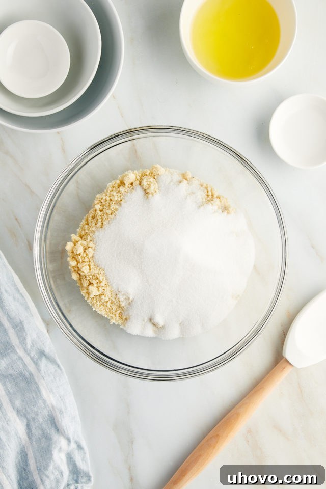 Overhead view of granulated sugar being added to almond flour in a mixing bowl, illustrating the first step of combining dry ingredients for Amaretti cookies.