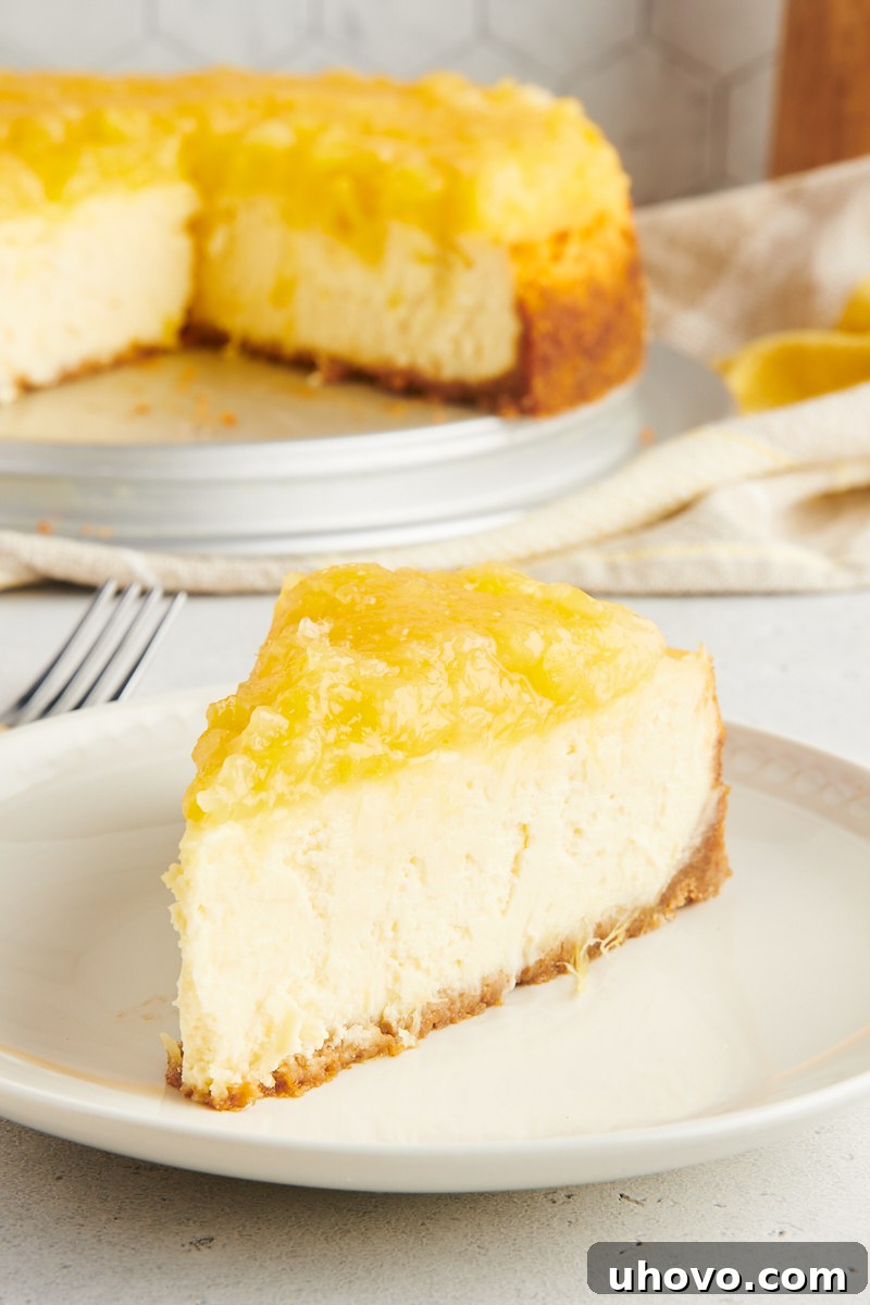 A close-up view of a single slice of pineapple cheesecake on a white plate in the foreground, with the remaining whole cheesecake, still in its springform pan, blurred in the background, creating a mouth-watering scene.
