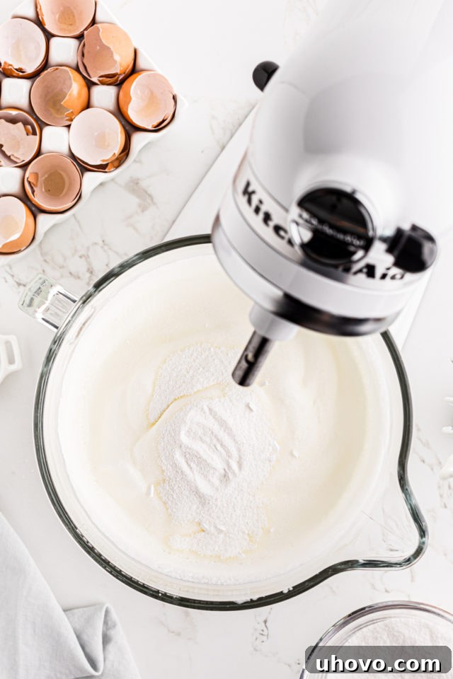 The remaining superfine sugar being gradually added to whipped egg whites in a stand mixer, creating a glossy, stiff meringue.