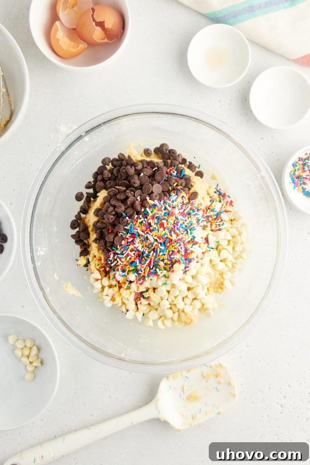 An overhead view of the finished cookie dough in a glass mixing bowl, with an array of semisweet chocolate chips and vibrant rainbow sprinkles having just been folded in, ready for chilling.