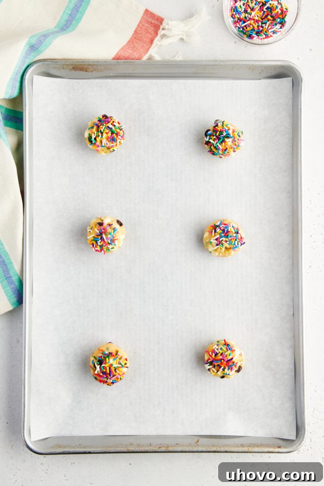 A baking sheet lined with parchment paper, neatly arranged with portioned balls of sprinkle chocolate chip cookie dough, some topped with extra sprinkles, spaced evenly for baking.