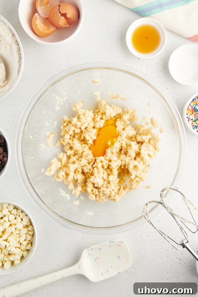 An overhead shot showing a single large egg being cracked and added to the creamy mixture of butter and sugars in a glass mixing bowl, mid-preparation for cookie dough.
