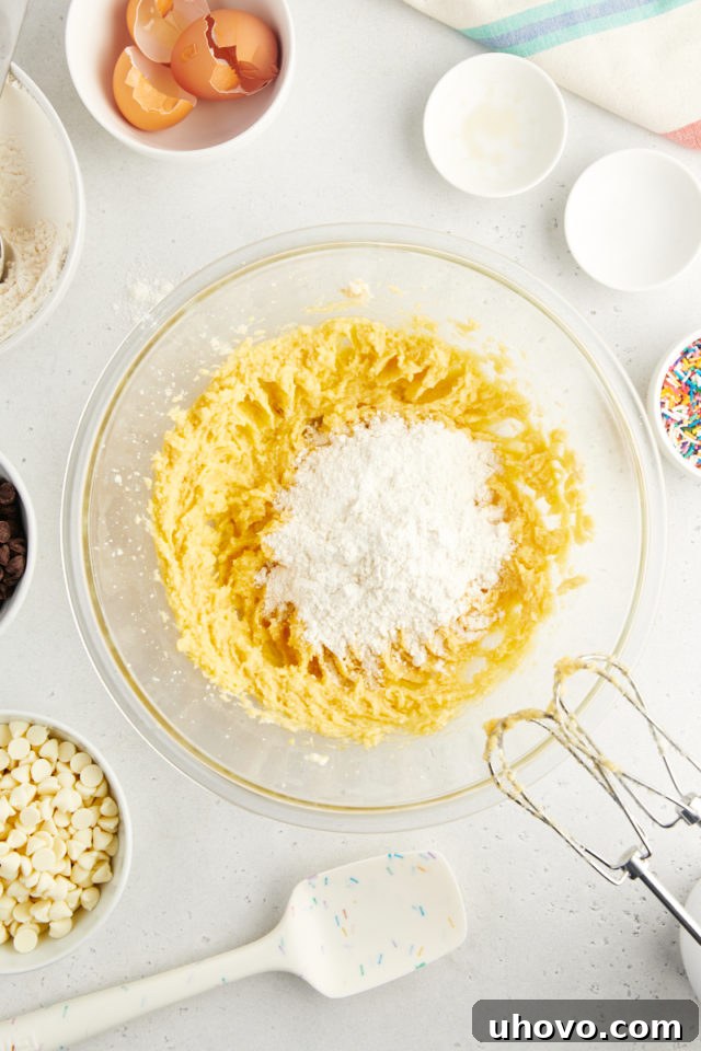 An overhead view showing the whisked dry ingredients (flour, cornstarch, baking soda, salt) being gradually added to the wet creamed mixture in a large glass mixing bowl, forming the cookie dough.