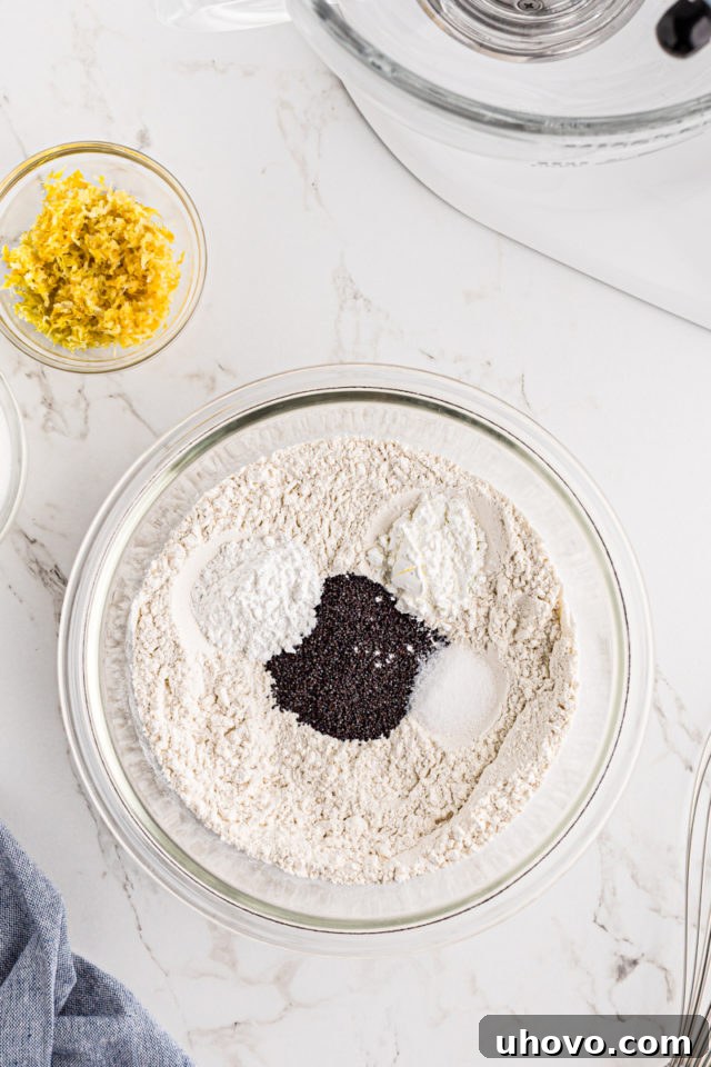 Overhead view of flour, baking powder, cornstarch, salt, and poppy seeds in a glass mixing bowl, ready to be whisked.
