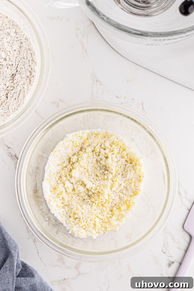 Overhead view of granulated sugar and fresh lemon zest mixed together in a glass mixing bowl, appearing crumbly and fragrant.