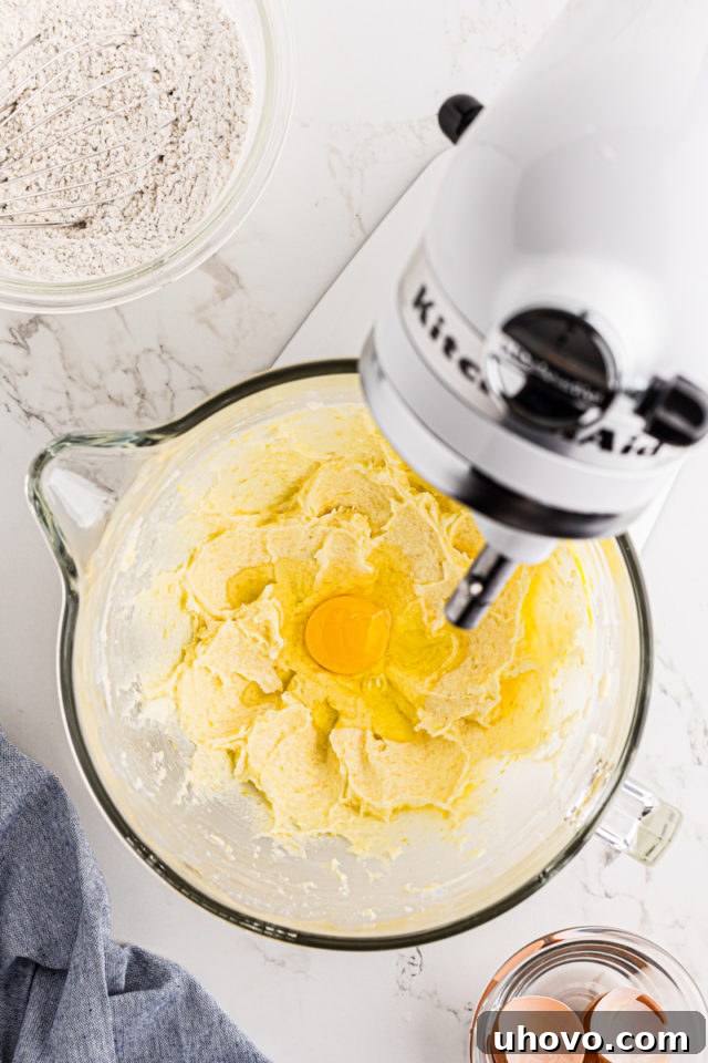 Overhead view of a cracked egg being added to the creamed butter and sugar mixture in a stand mixer.