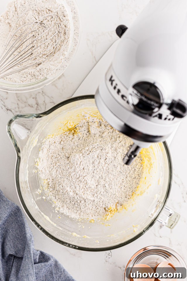 Overhead view of the dry flour mixture being gradually added to the wet ingredients in a stand mixer to form cookie dough.