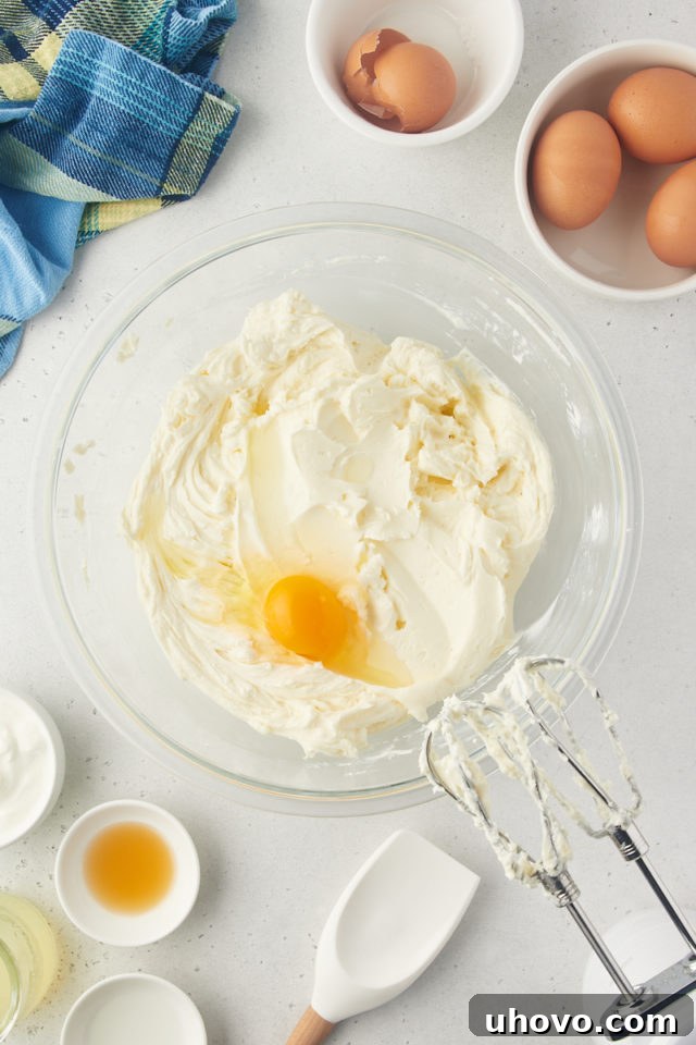 Overhead view of an egg being added to the creamy lemon cheesecake mixture in a glass bowl.