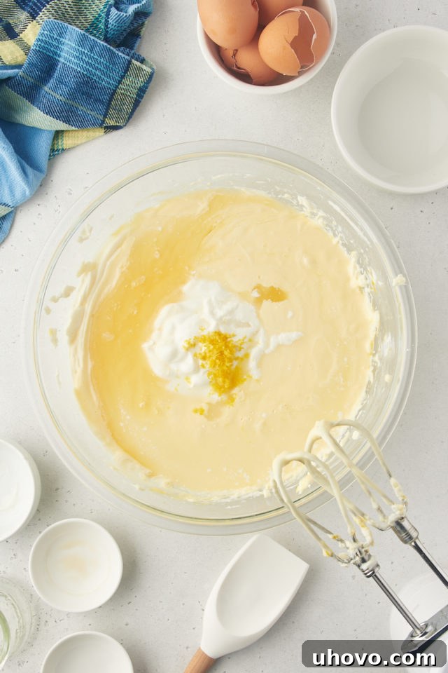 Overhead view of sour cream, lemon juice, lemon zest, vanilla extract, and lemon extract being added to the cheesecake batter.