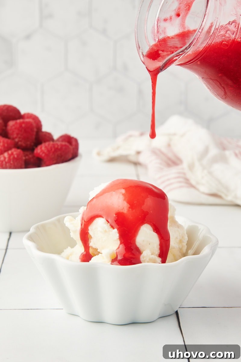Vibrant raspberry coulis being drizzled generously over a scoop of vanilla ice cream in a white bowl.