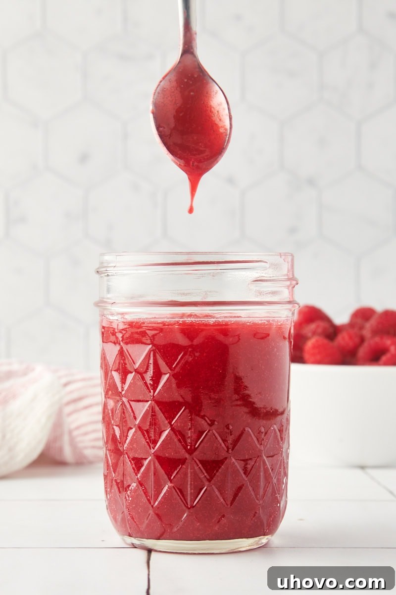 A spoonful of vibrant raspberry coulis lifted from a glass jar, showcasing its smooth texture.
