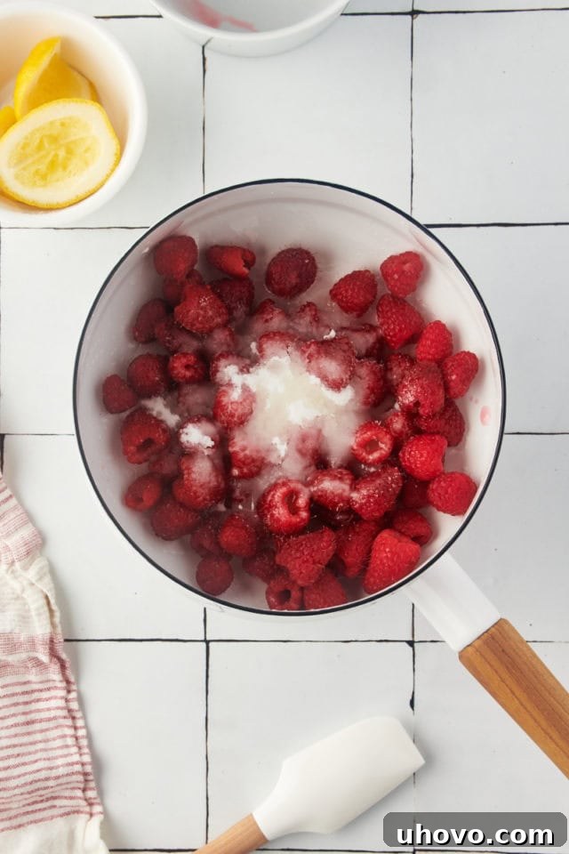 Overhead view of raspberries, sugar, and lemon juice combined in a saucepan, ready for cooking.