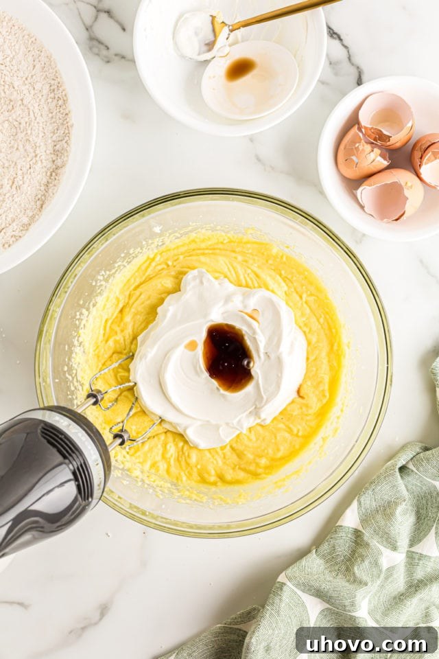 Overhead view of an electric mixer blending room temperature sour cream and vanilla extract into the cake batter base.