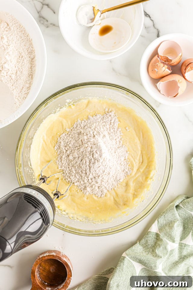 Overhead view of dry flour mixture being added to the wet cake batter in a glass bowl with an electric mixer, in preparation for final mixing.