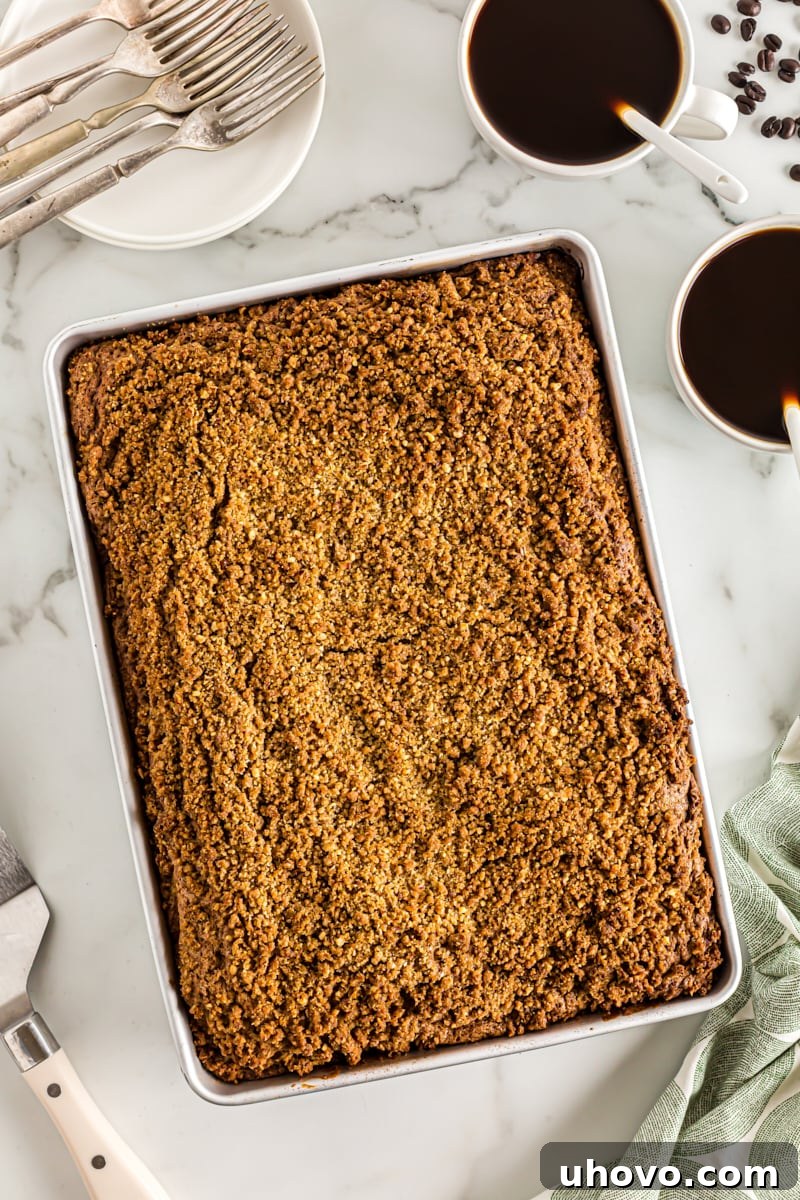 Overhead view of a freshly baked sour cream coffee cake in its pan, accompanied by two steaming cups of black coffee, ready for enjoyment.