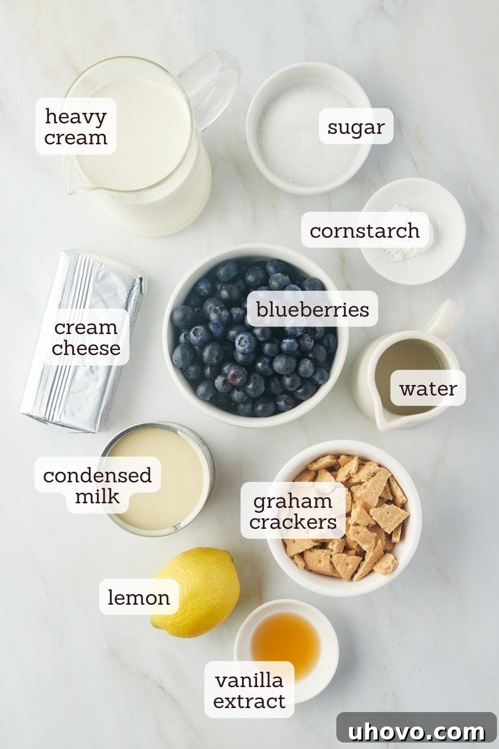 Overhead view of all the fresh ingredients for homemade Blueberry Cheesecake Ice Cream laid out on a white surface, including blueberries, cream cheese, sweetened condensed milk, heavy cream, vanilla, sugar, cornstarch, and graham crackers.