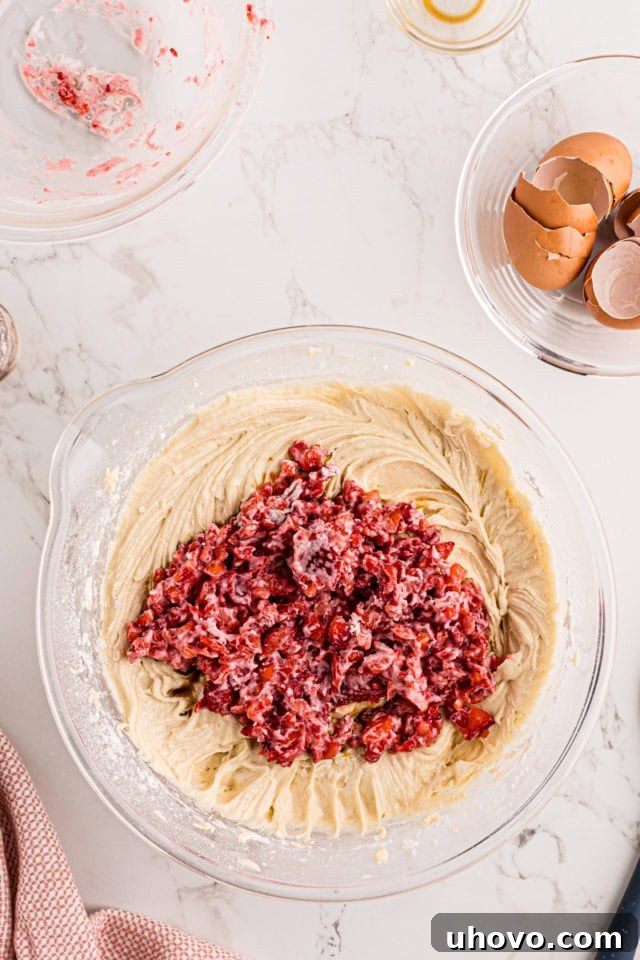 A close-up overhead shot showing chopped strawberries, dusted lightly with flour, being carefully added to the strawberry pound cake batter in a large glass mixing bowl.