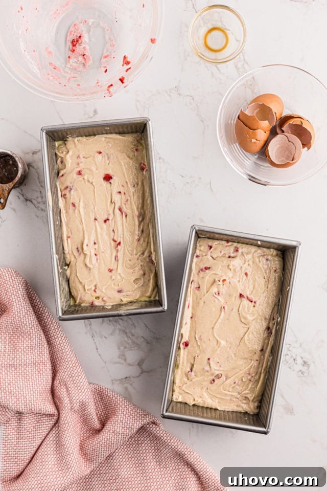 Overhead perspective of two loaf pans, evenly filled with strawberry pound cake batter, ready for baking. The batter is speckled with red strawberry pieces.