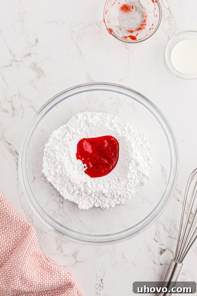 An overhead shot showcasing the ingredients for the strawberry glaze: confectioners' sugar, freshly pureed strawberries, and milk, all in a clear glass mixing bowl, ready to be combined.