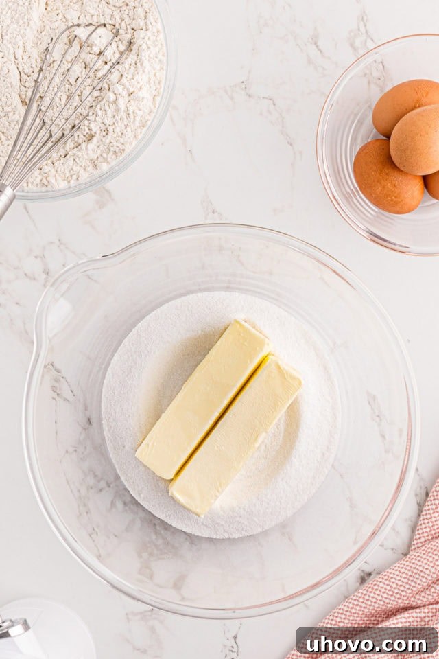 A close-up, overhead view of softened unsalted butter and granulated sugar in a clear glass mixing bowl, being creamed together to form a light and fluffy mixture.