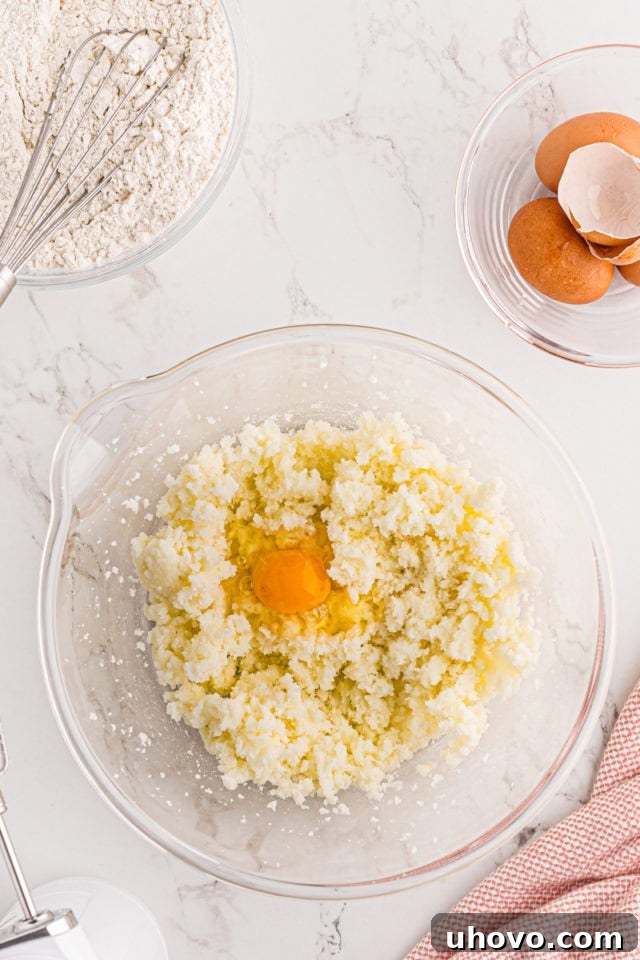 An overhead shot showing a single egg being cracked and added to the creamed butter and sugar mixture in a glass mixing bowl, highlighting the gradual incorporation of ingredients.