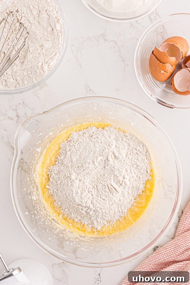 An overhead view capturing dry flour mixture being carefully added to the wet ingredients in a glass mixing bowl, illustrating the gradual incorporation process for the strawberry pound cake batter.